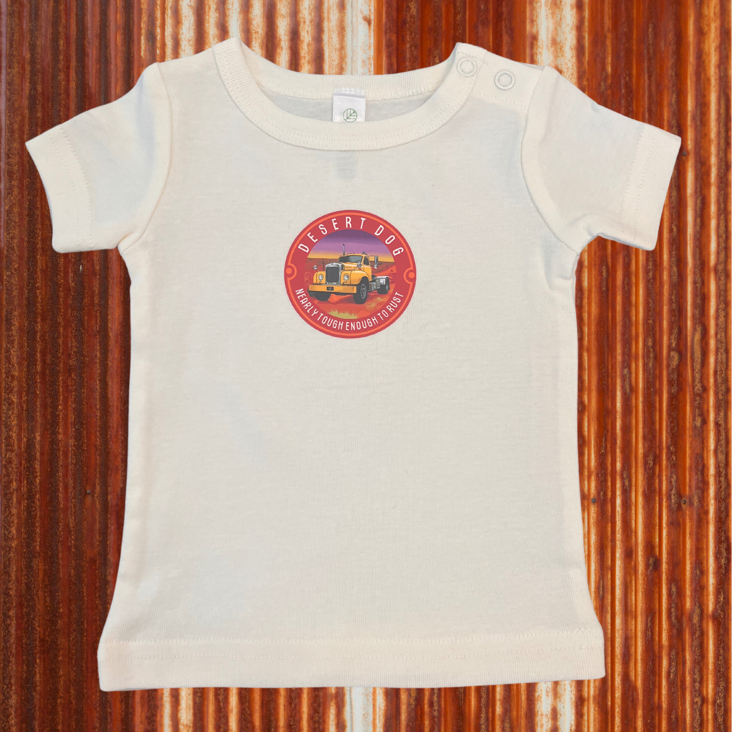 A cream coloured tiny tee with a B model Mack truck on the front, displayed on a rusty tin shed background.