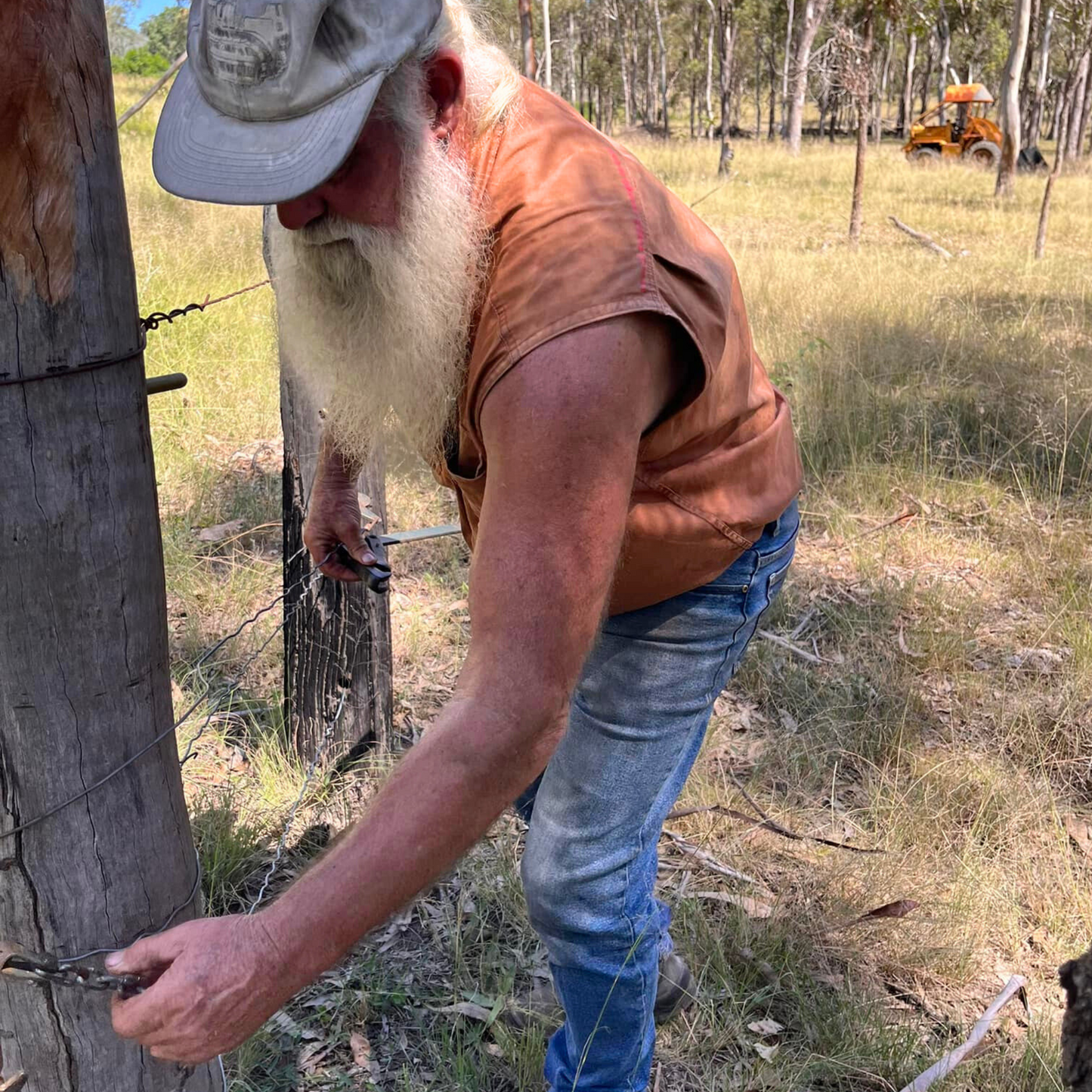 A bearded man wearing an old cap and a Desert Dog WorkTough shirt checking a farm fence with a loader in the background