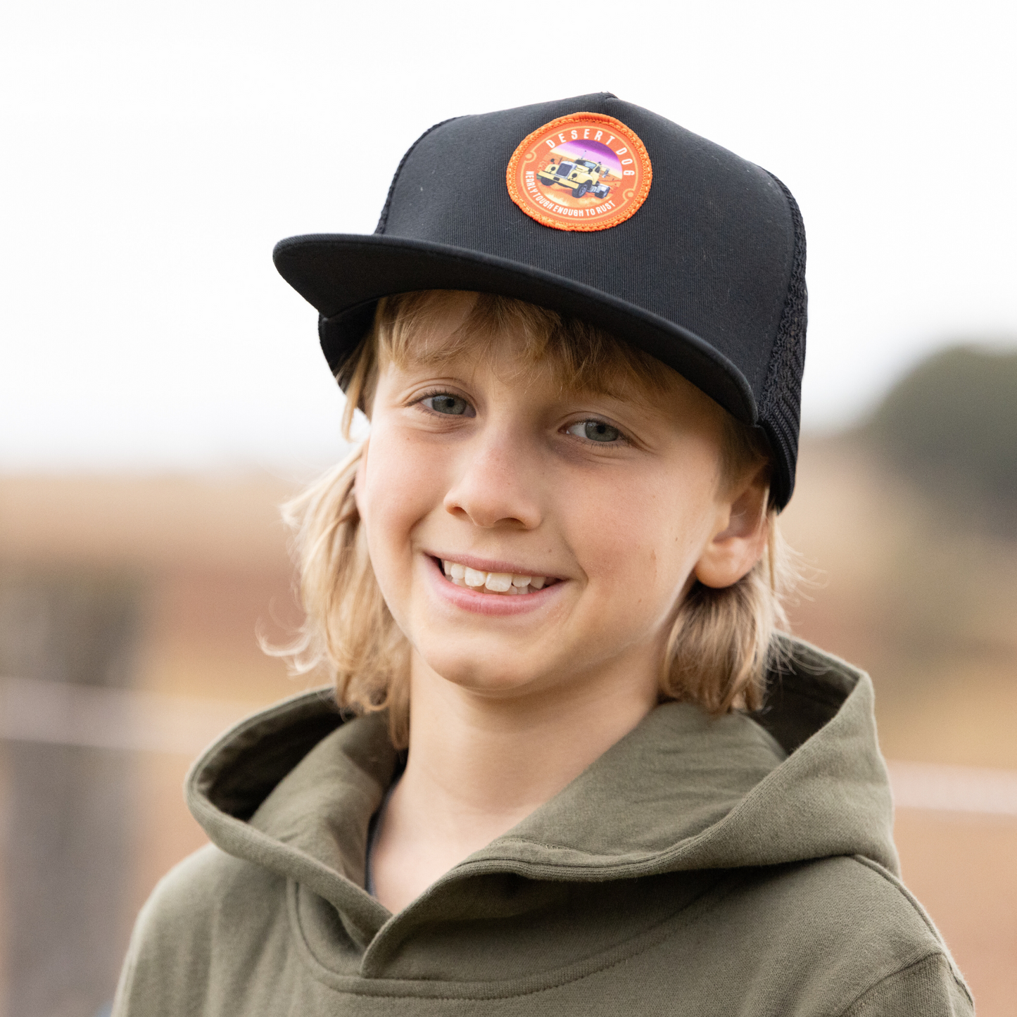 A smiling boy is wearing a black, flat peak trucker cap with a Desert Dog logo