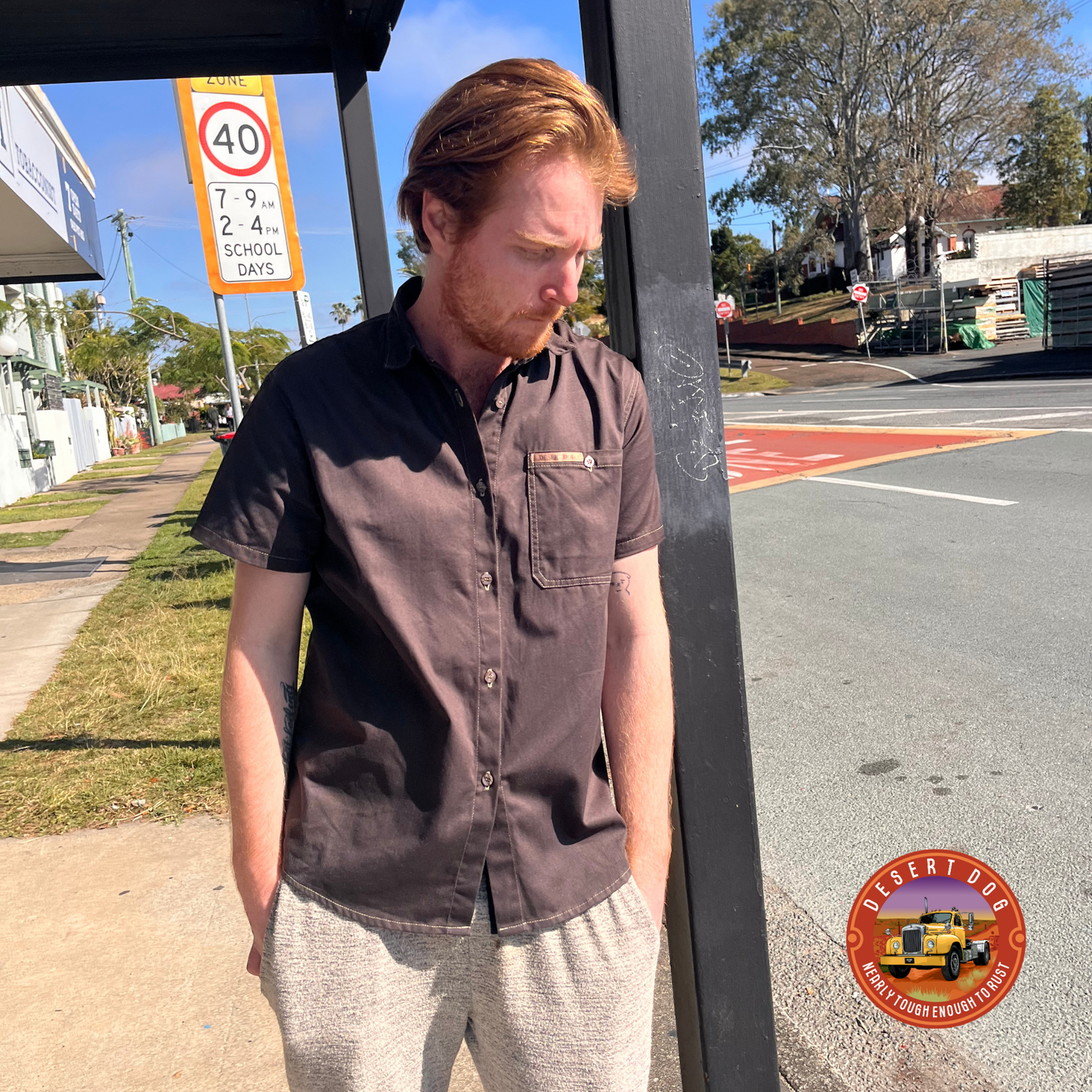 A red haired man leaning against a shop veranda post beside a road and wearing a dark brown Desert Dog WorkTough shirt with short sleeves.