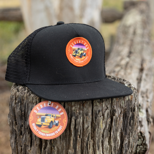 A blck trucker cap with a flat peak and Desert Dog logo  perched on a timber fence post