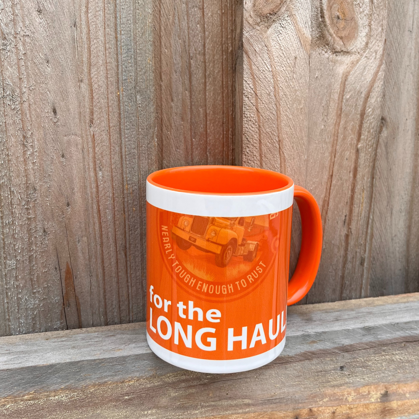 An orange and white coffee mug on a wooden fence featuring the Desert Dog B model Mack truck logo