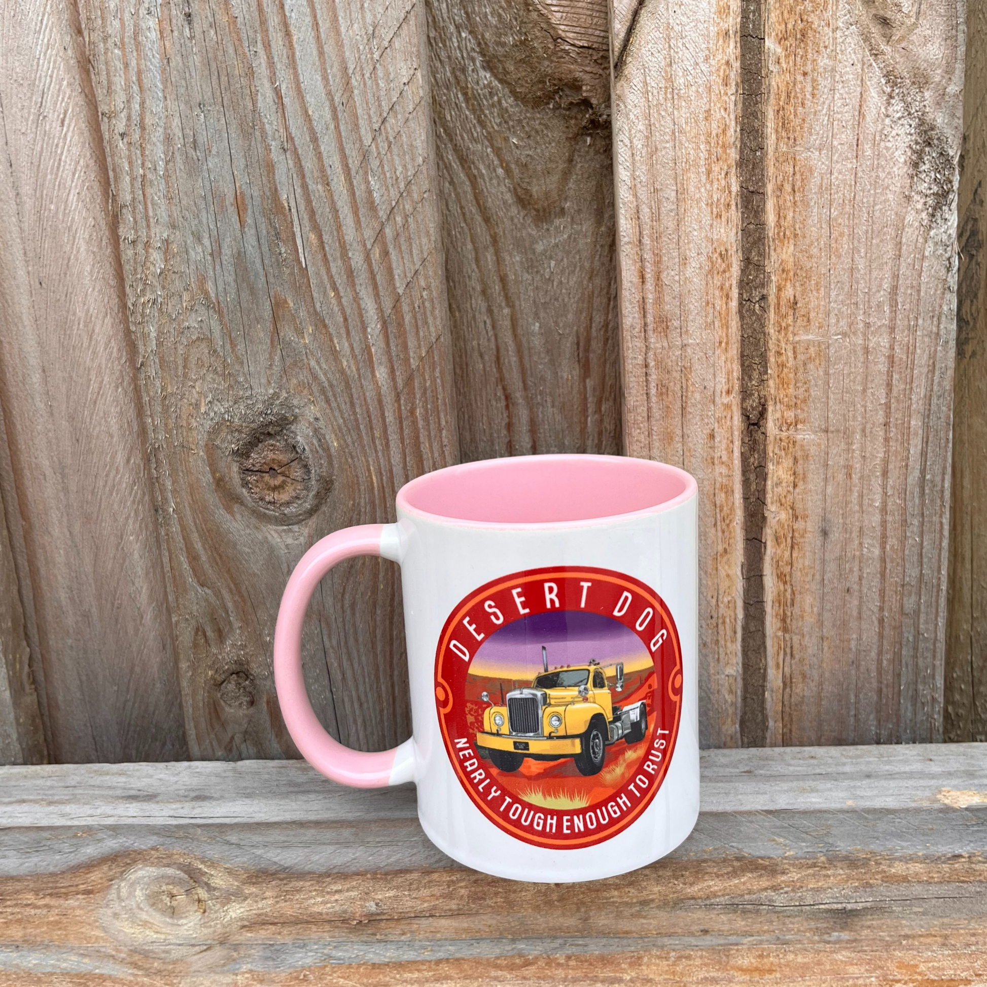 A pink and white coffee mug on a wooden fence featuring the Desert Dog B model Mack truck logo