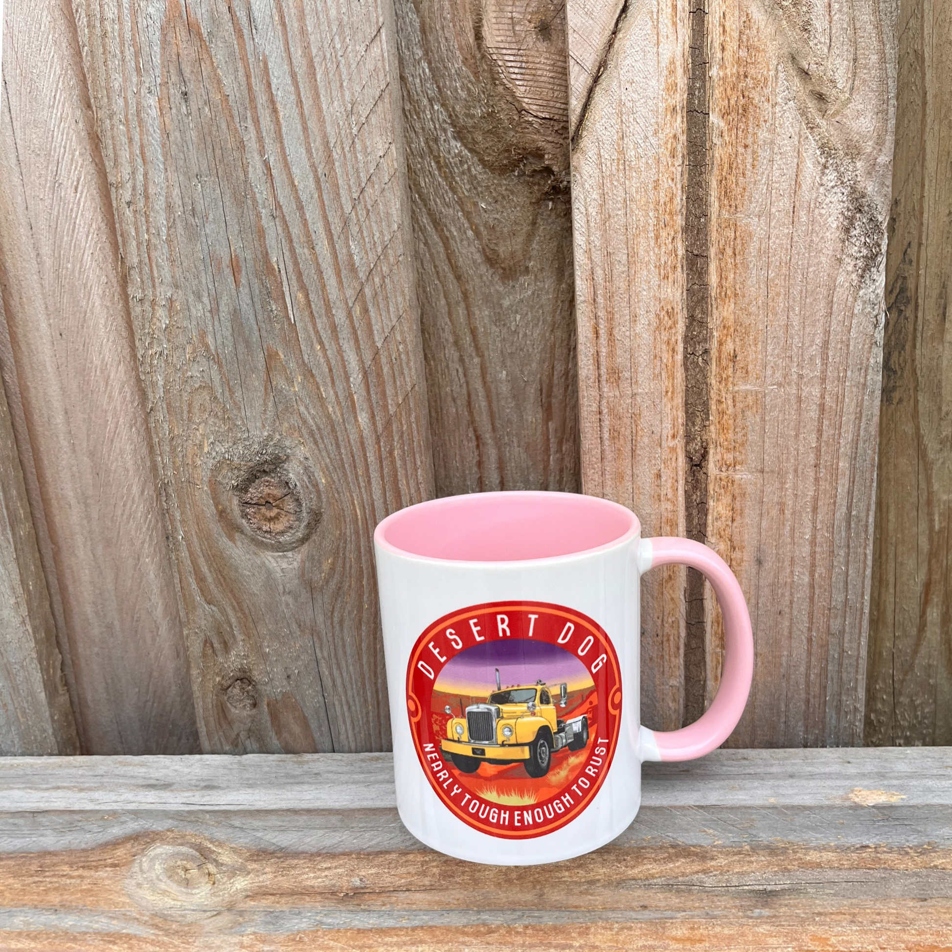 A pink and white coffee mug on a wooden fence featuring the Desert Dog B model Mack truck logo