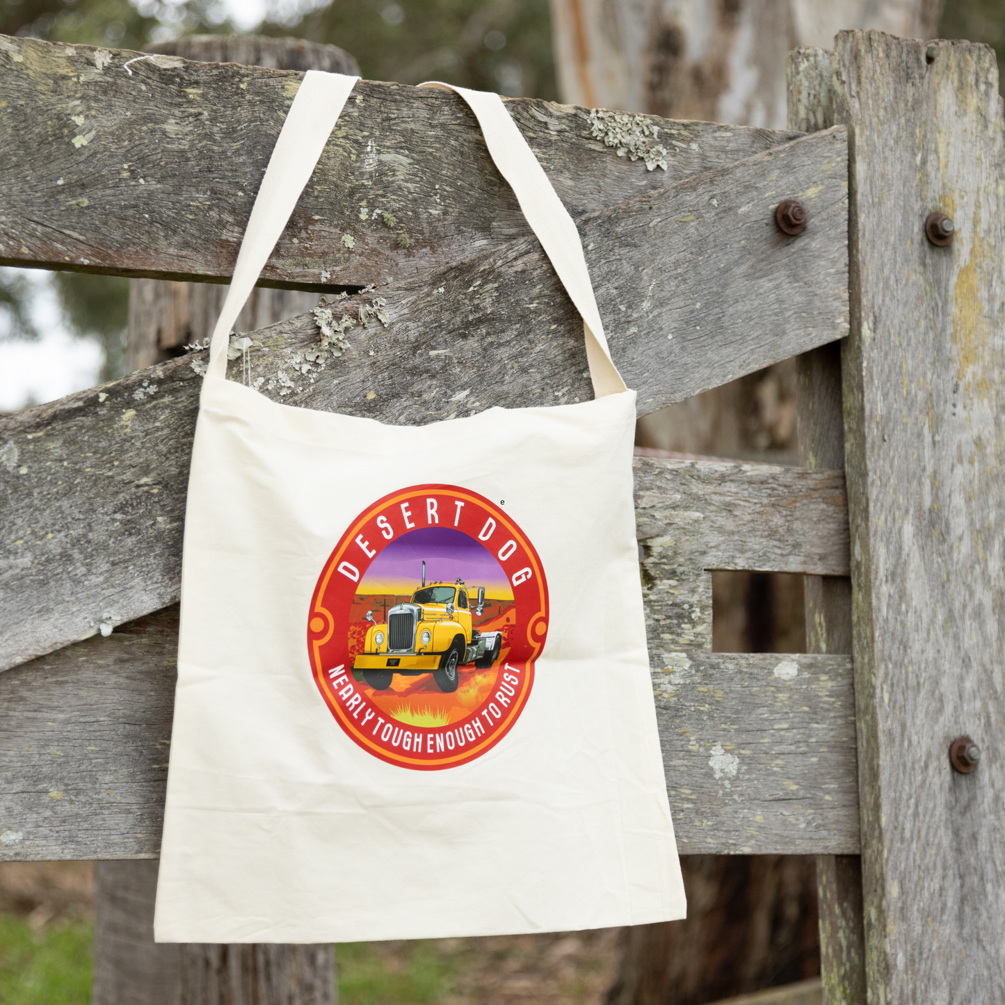 A calico tote bag with a Desert Dog logo hanging on an old wooden gate