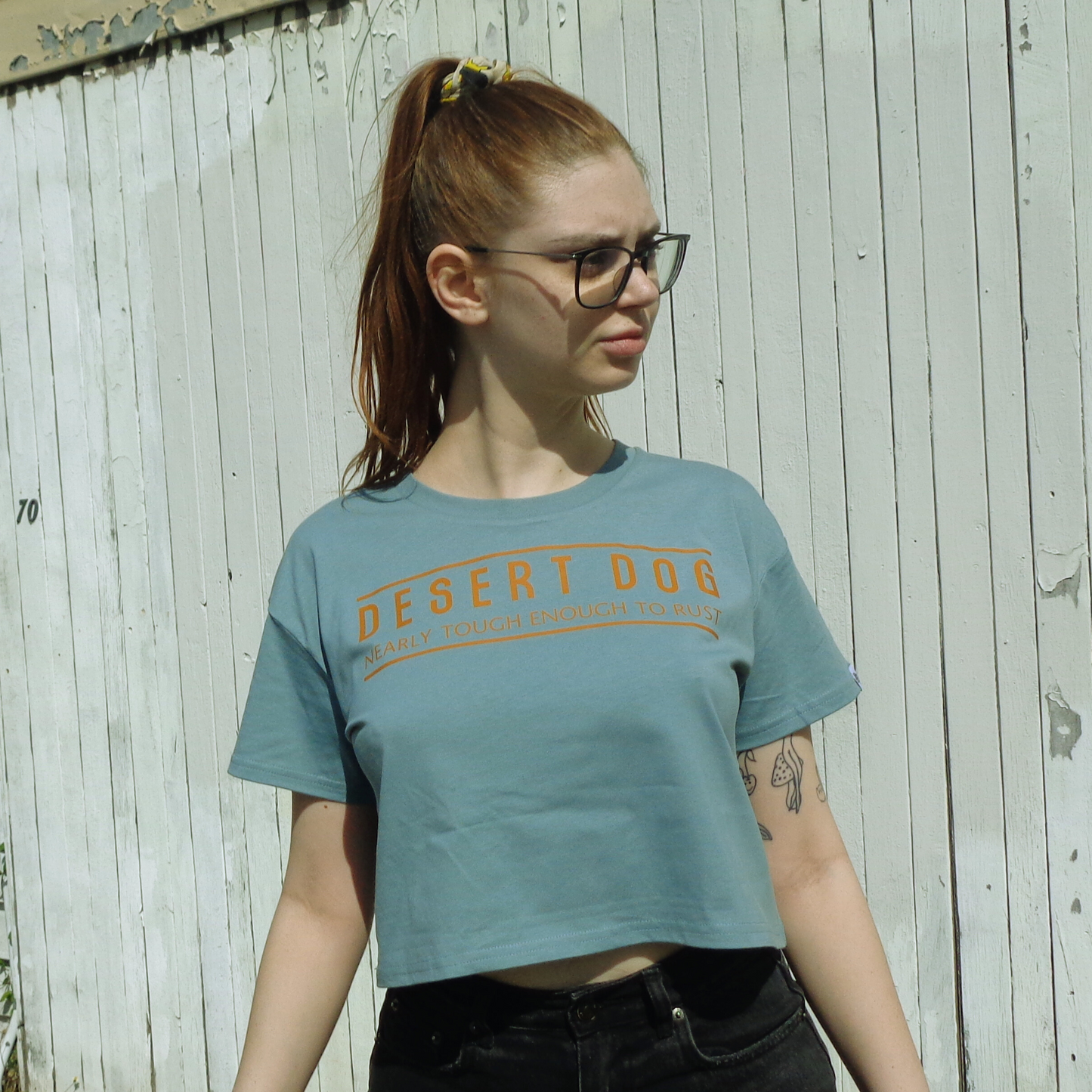 Daphne is wearing a mineral coloured Desert Dog crop tee and standing in front of an old building. She has red hair in a ponytail and is wearing glasses.
