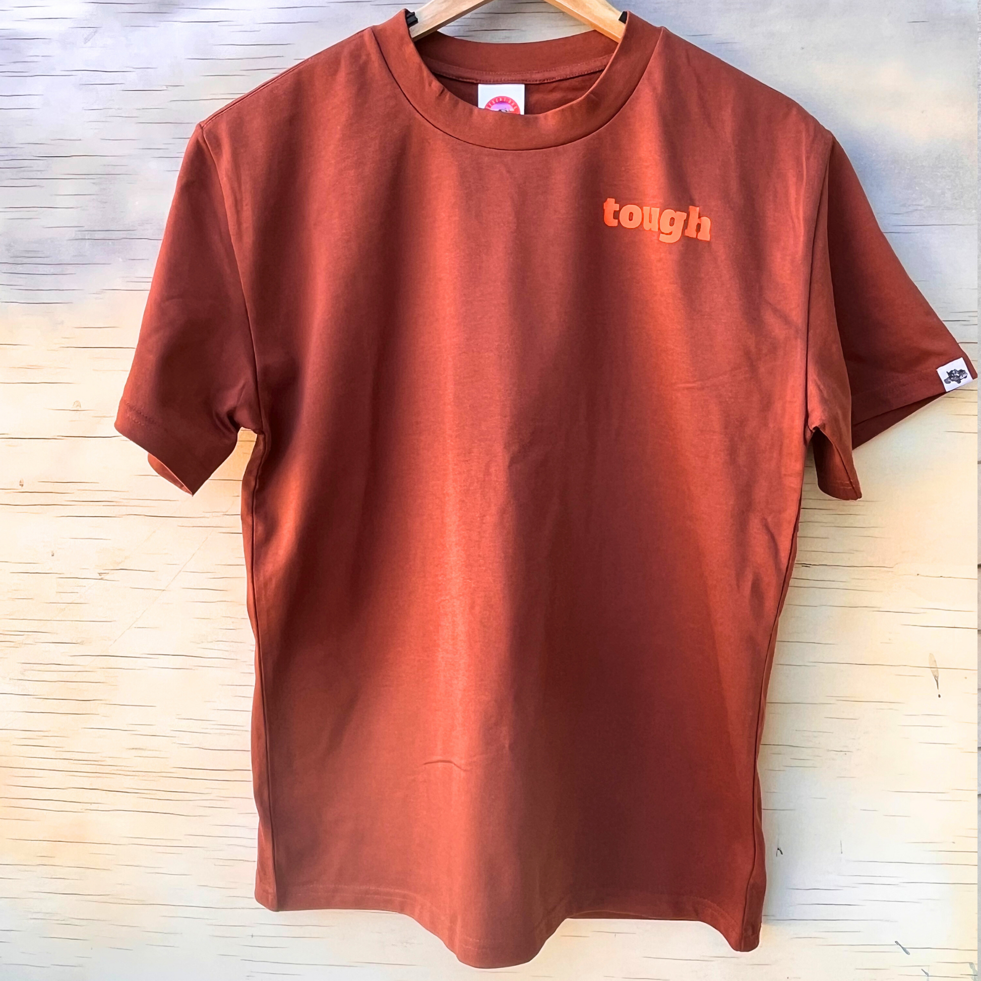 The front of a men's rust coloured t-shirt, showing the word "tough" in orange text on the left chest area. A B model Mack truck is tagged onto the left sleeve.