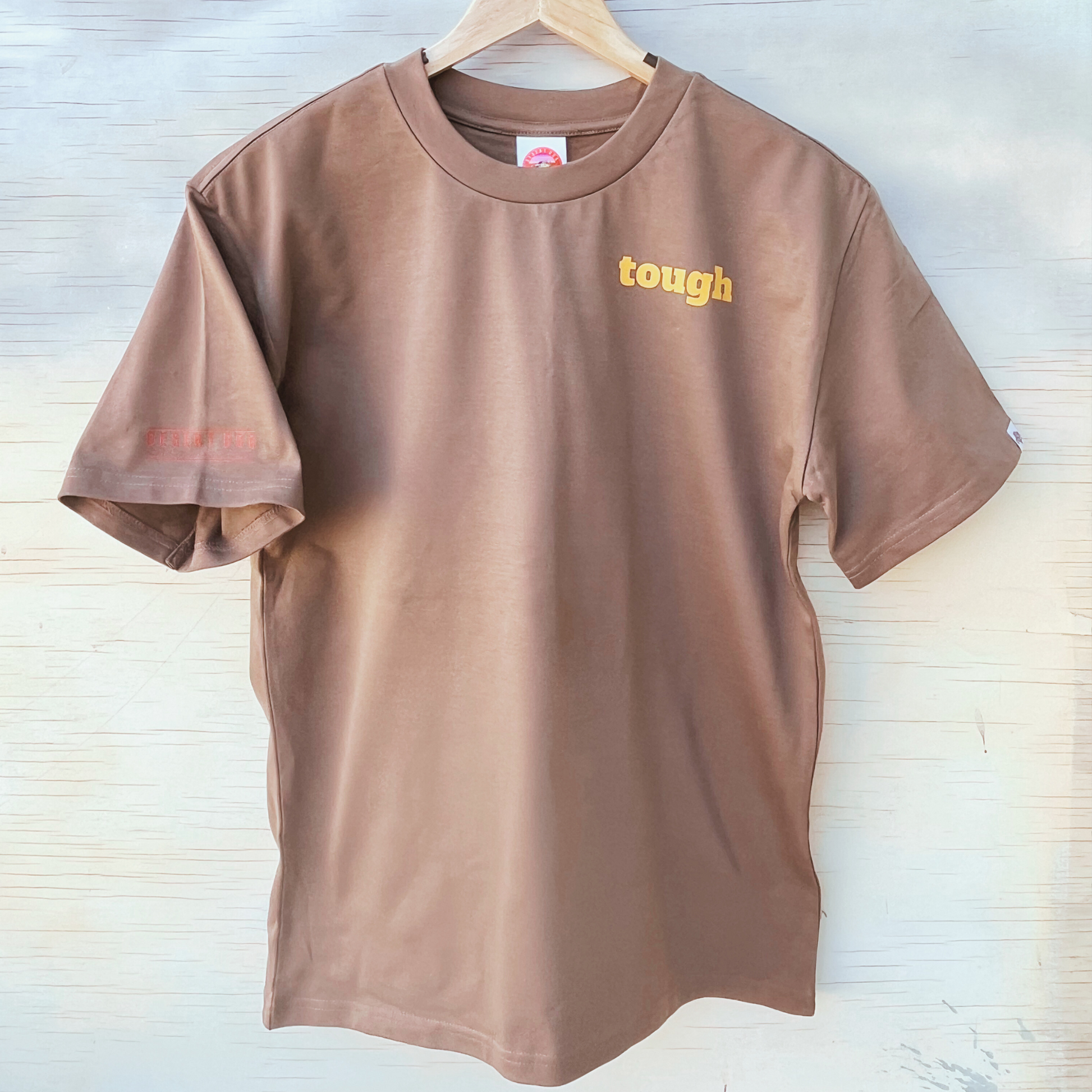 The front of a men's walnut coloured t-shirt, showing the word "tough" in yellow text on the left chest area. A B model Mack truck is tagged onto the left sleeve.