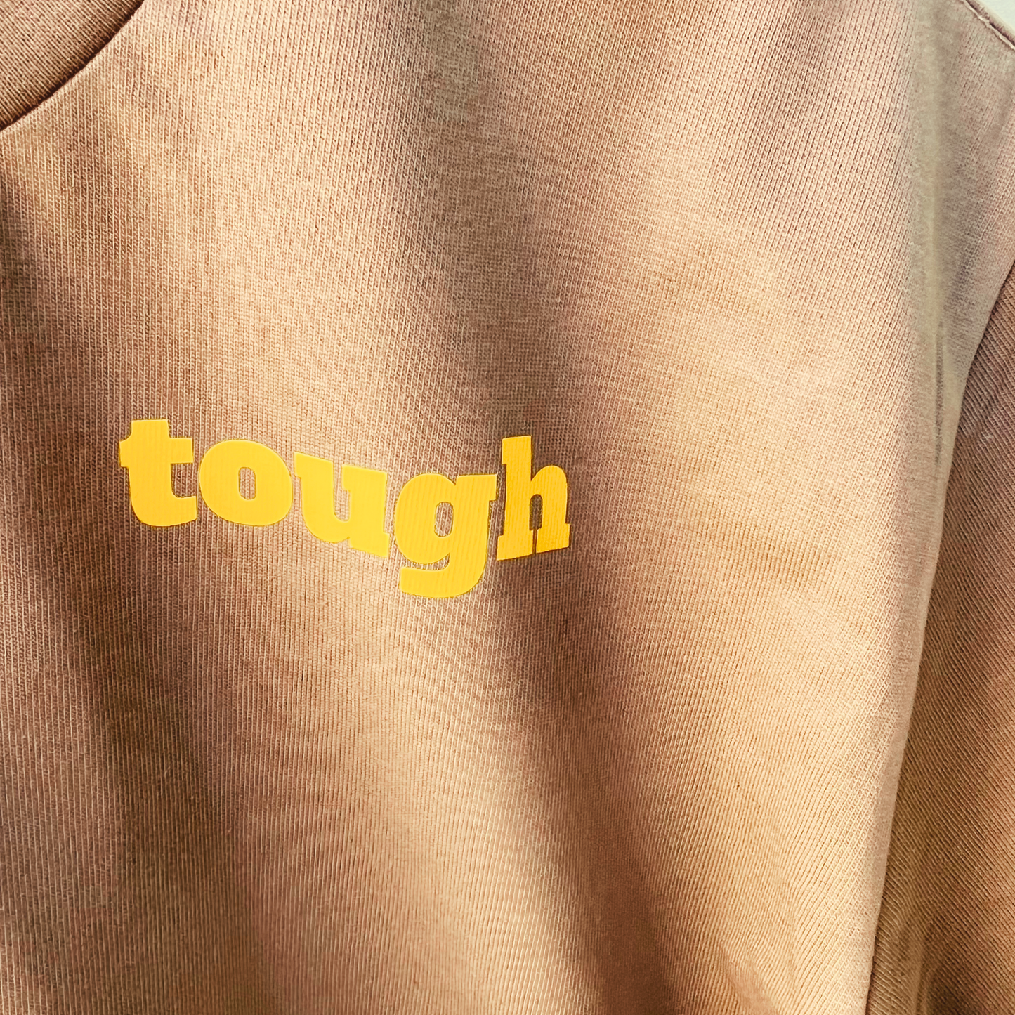 A close up detail of a men's walnut coloured t-shirt with the word "tough" printed in yellow text.