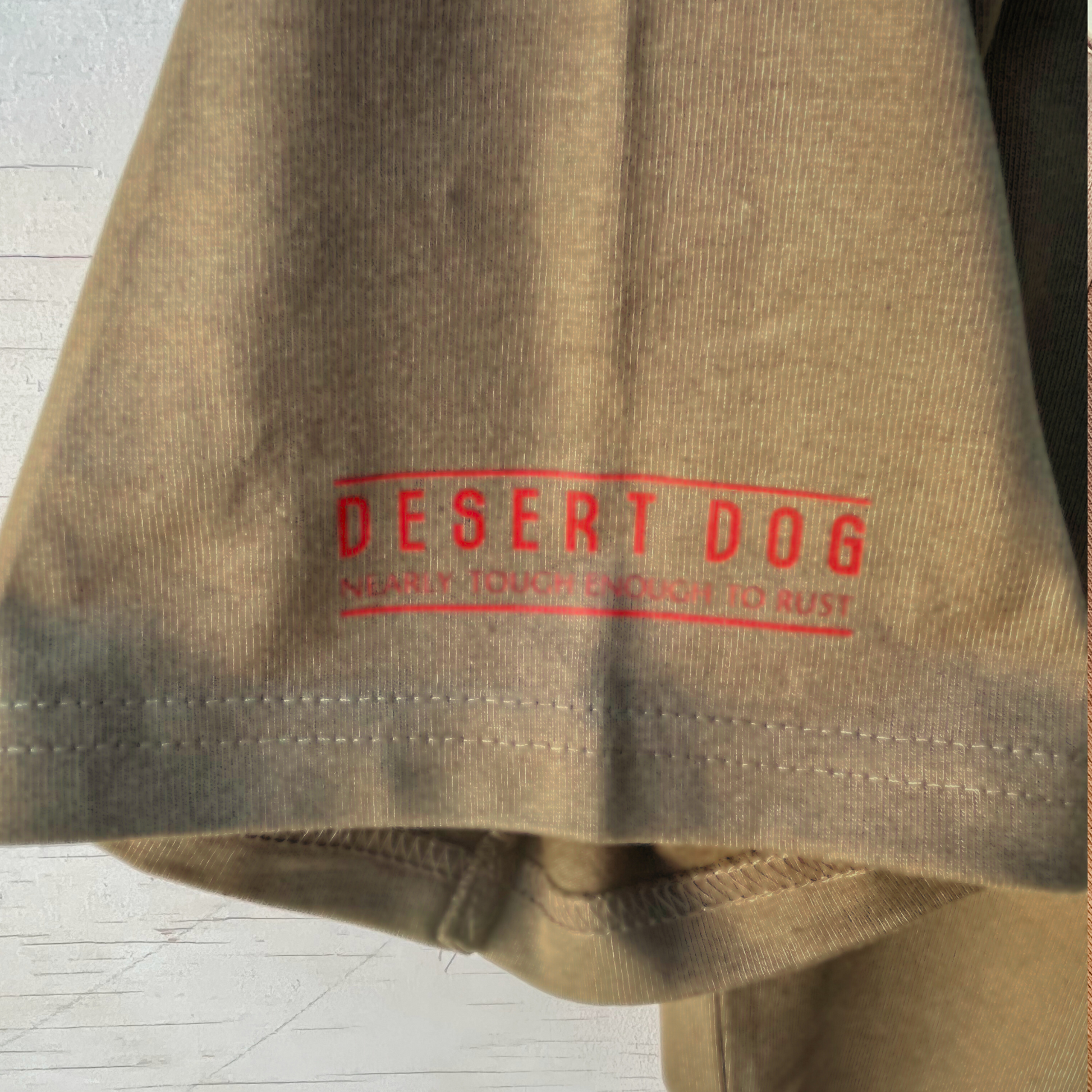 A close up detail of a men's walnut coloured t-shirt sleeve showing the Desert Dog text logo and the words "nearly tough enough to rust".