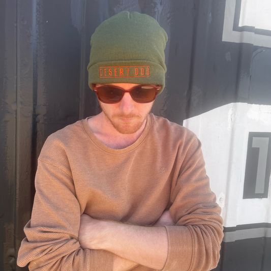 A male model is leaning against the wall of a tattoo shop wearing an army green beanie with the words Desert Dog embroidered on the cuff. He is wearing sunglasses and has his arms folded.