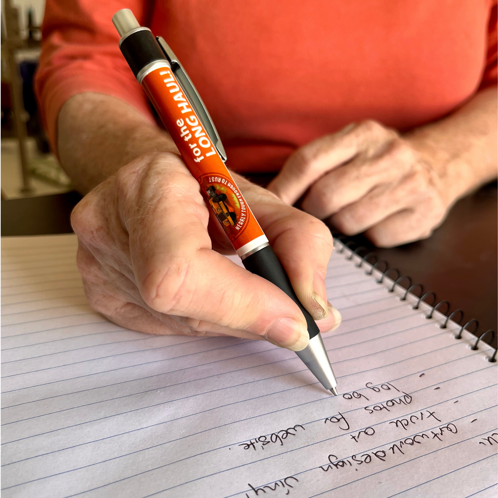 A push button pen featuring the Desert Dog logo and the words "for the long haul". The woman holding the pen is writing on a spiral note pad.