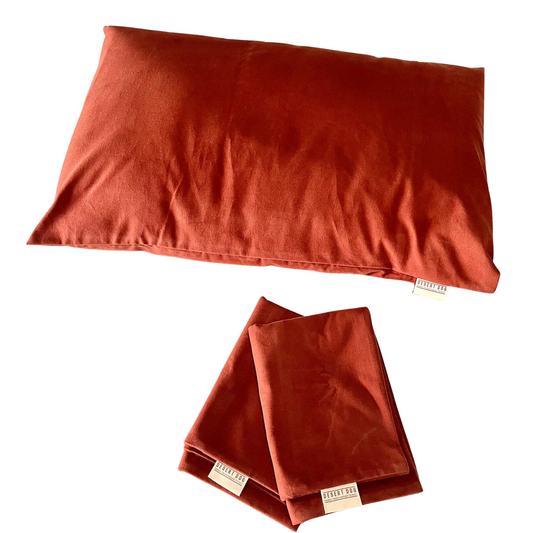 A pillow with a rust coloured pillow case and a pair of folded  rust coloured pillow cases