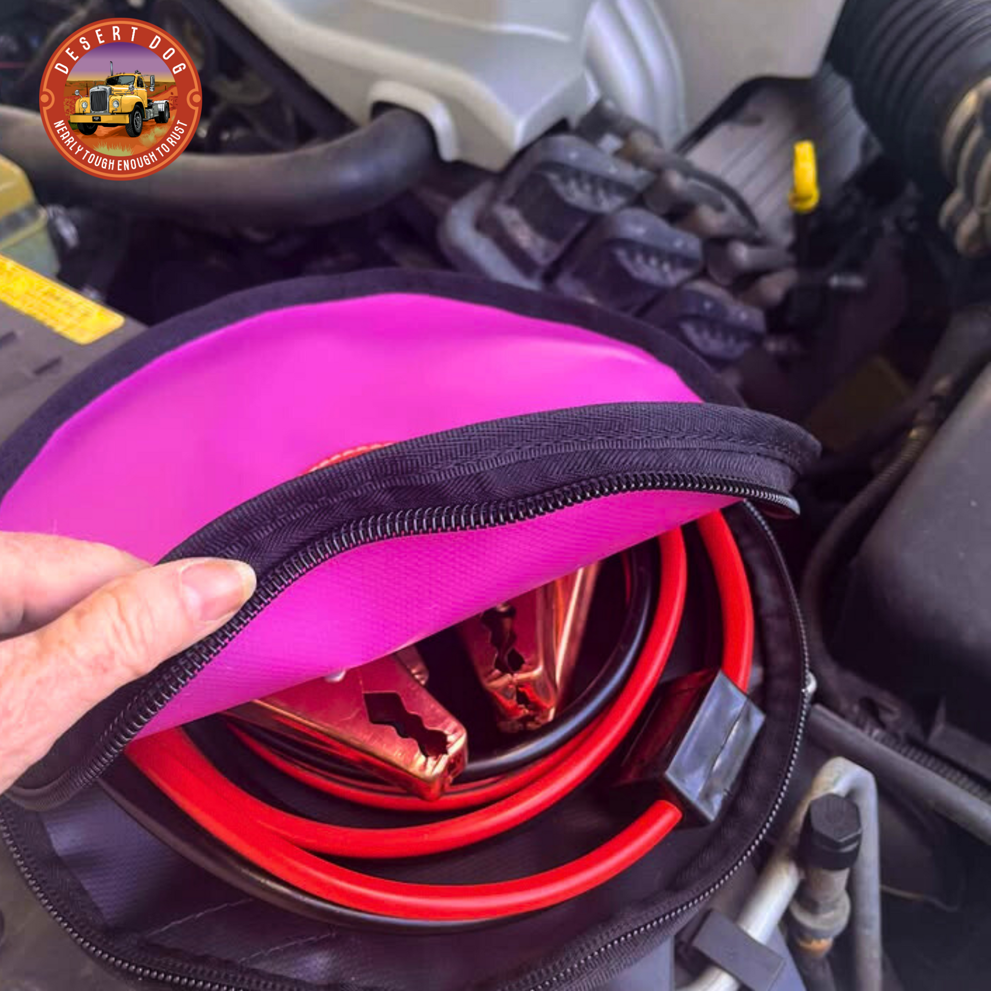 Pink 30cm Desert Dog rope bag with jump leads coiled inside and visible through the opened zip of the bag. The bag is sitting in the engine bay of a V6 holden 