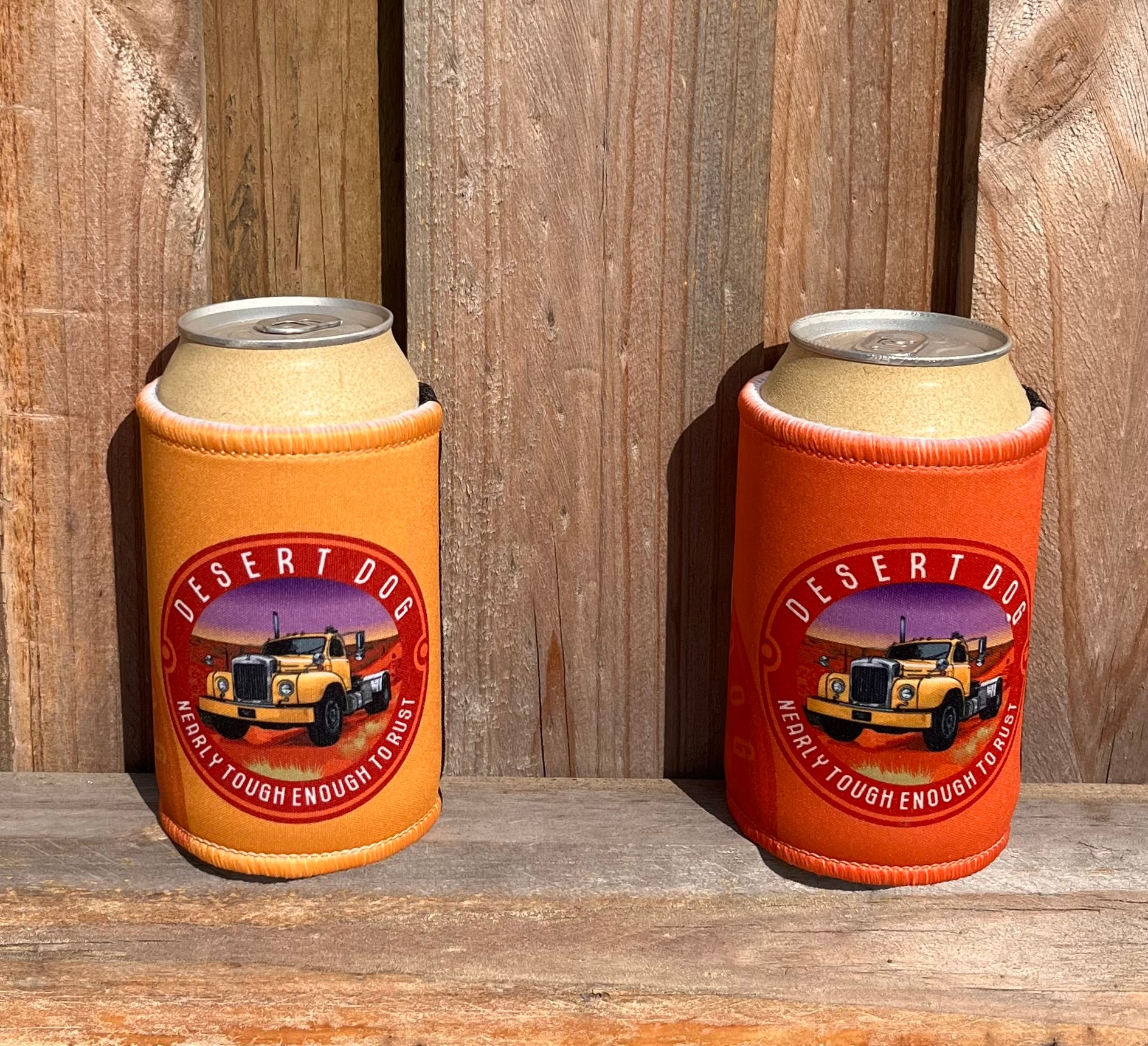 A pair of signature stubby holders, one orange and one yellow, with the Desert Dog logo of a B model Mack truck sitting on a wooden fence