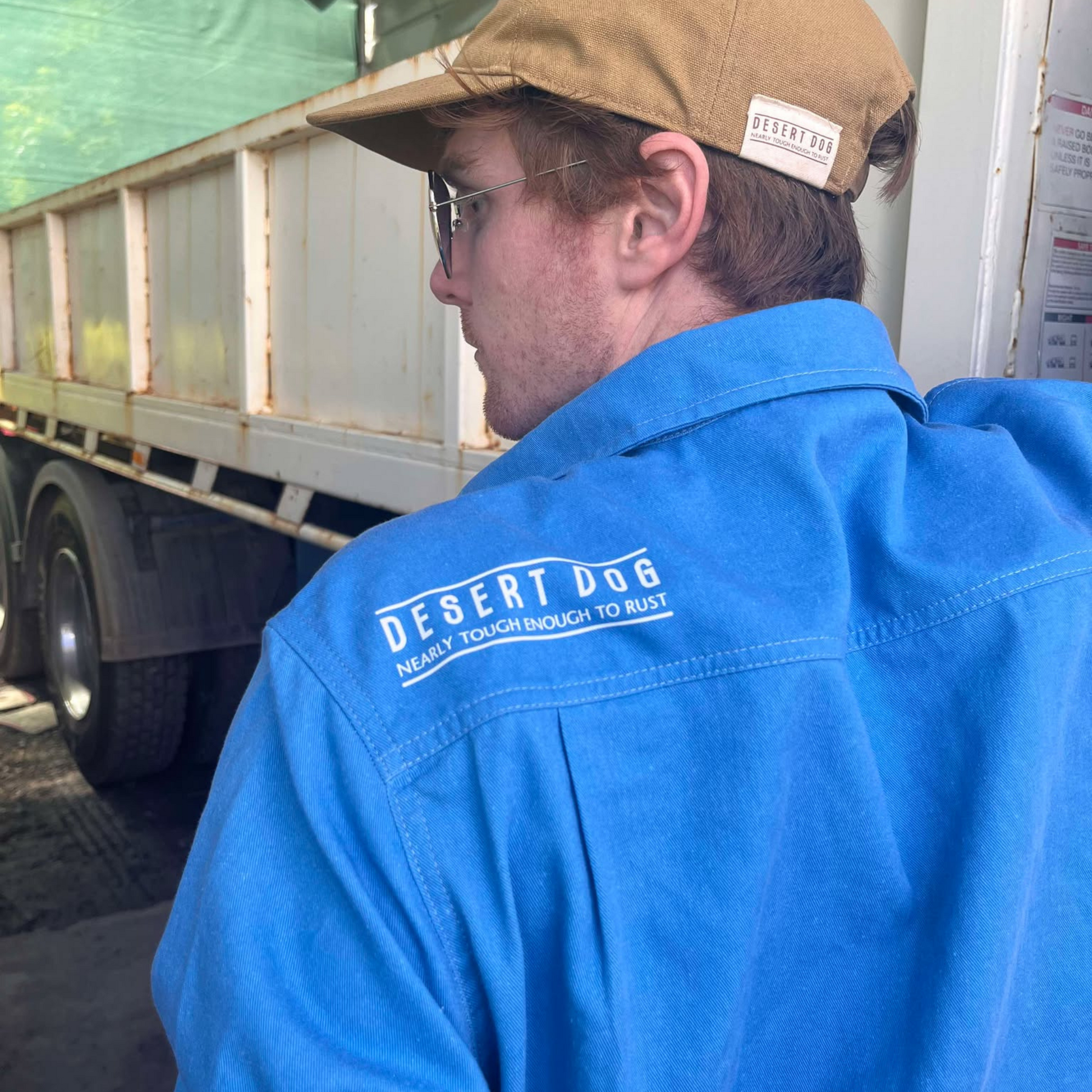 A truck driver standing beside his truck wearing a blue Desert Dog WorkTough shirt showing the logo on the back yoke. Also wearing a cap and sunglasses.