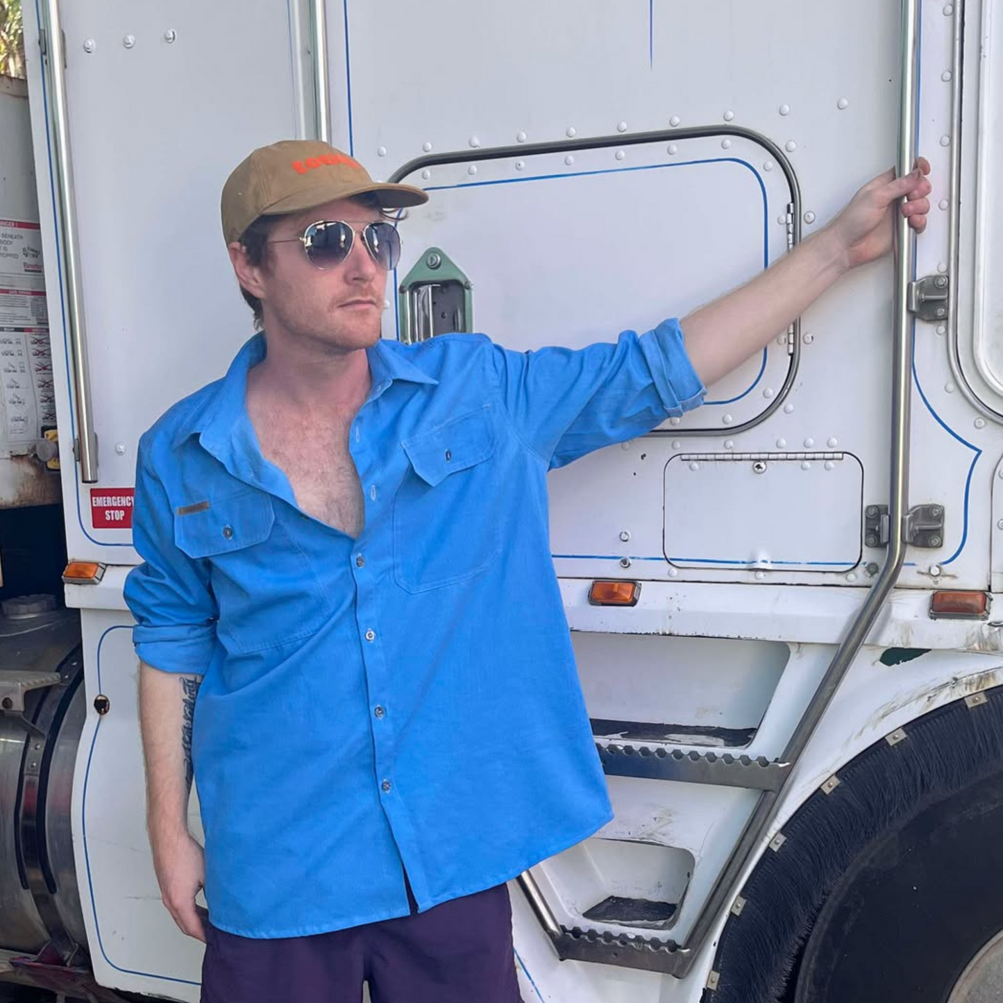 Male truck driver standing beside the steps of a prime mover wearing a blue Desert Dog WorkTough shirt with long sleeves, a cap and sungalsses