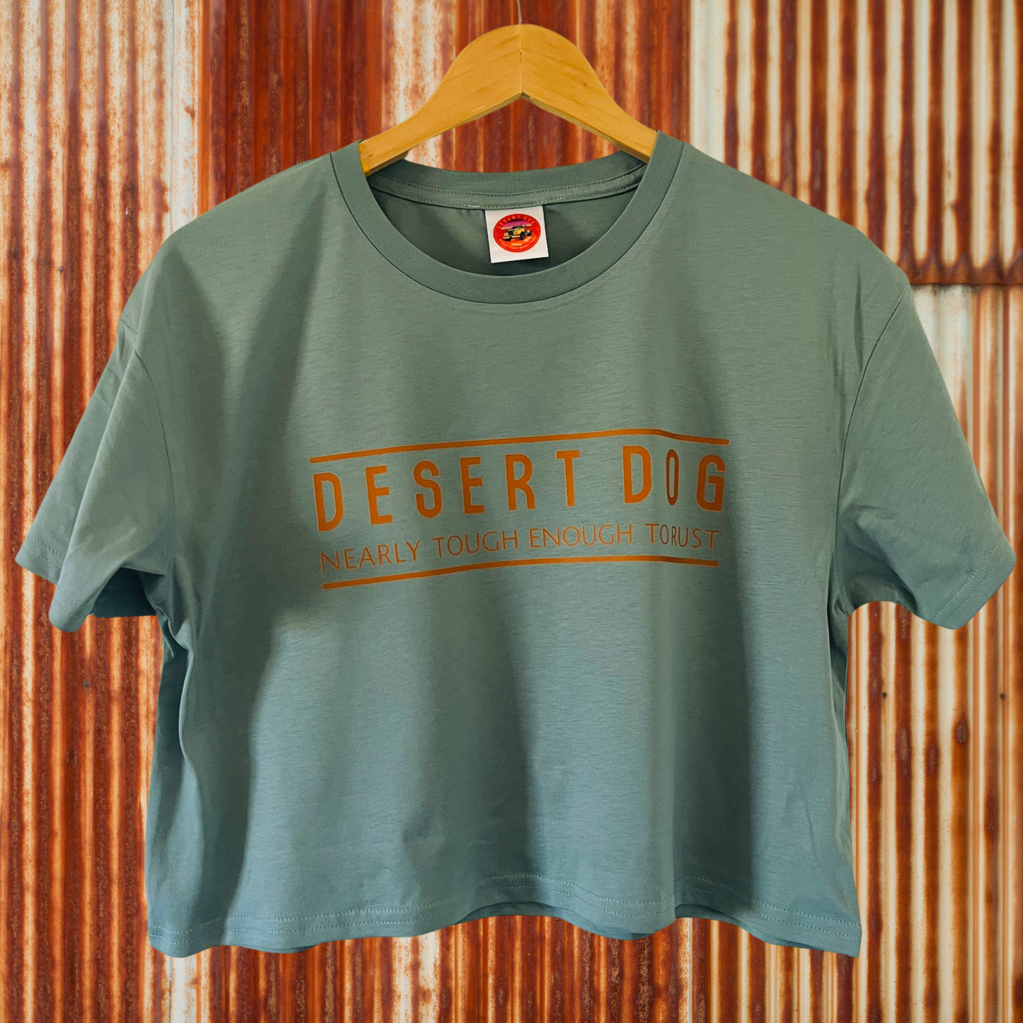 a mineral coloured ladies crop top with the Desert Dog text logo printed on the front. The tee is hanging against a rusty corrugated iron wall.