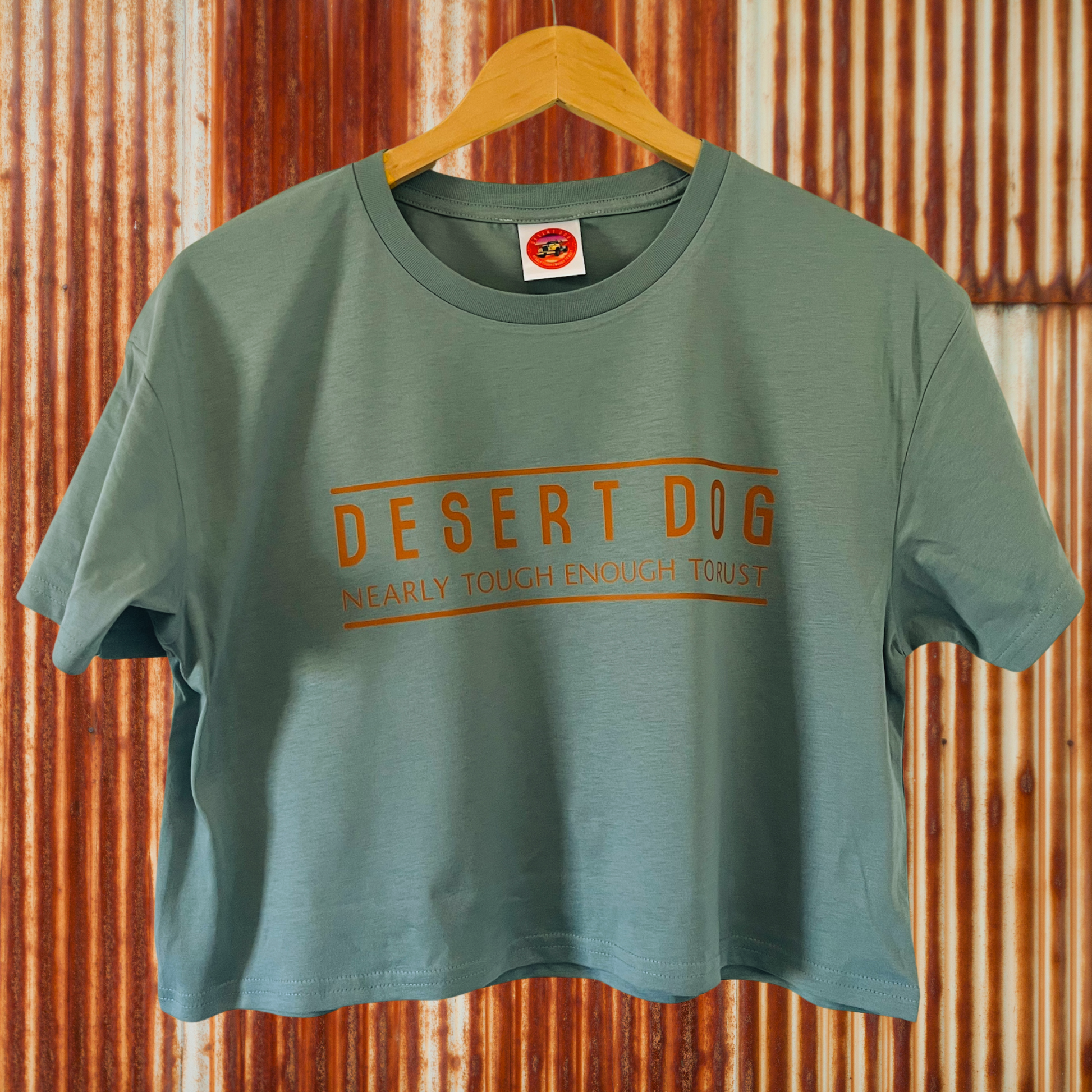 a mineral coloured ladies crop top with the Desert Dog text logo printed on the front. The tee is hanging against a rusty corrugated iron wall.